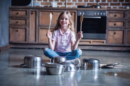 Foto de Linda niña está sosteniendo cucharas de madera, mirando a la cámara y sonriendo mientras toca la batería con los platos en la cocina - Imagen libre de derechos