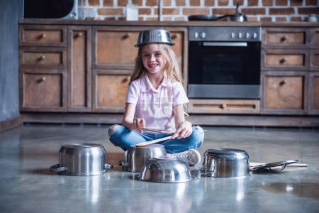 Foto de Linda niña está utilizando cucharas de madera, mirando a la cámara y sonriendo mientras toca la batería con platos en la cocina - Imagen libre de derechos