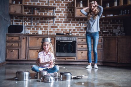 Photo for Cute little girl is using wooden spoons, looking at camera and smiling while playing drums with dishes in kitchen, mom is clutching her head - Royalty Free Image