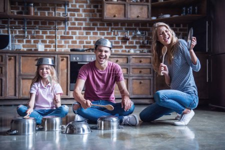 Foto de Linda niña y sus padres jóvenes están usando cucharas de madera y sonriendo mientras tocan la batería con platos en la cocina - Imagen libre de derechos