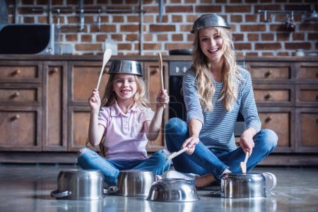 Foto de Linda niña y su madre están utilizando cucharas de madera, mirando a la cámara y sonriendo mientras toca la batería con platos en la cocina - Imagen libre de derechos