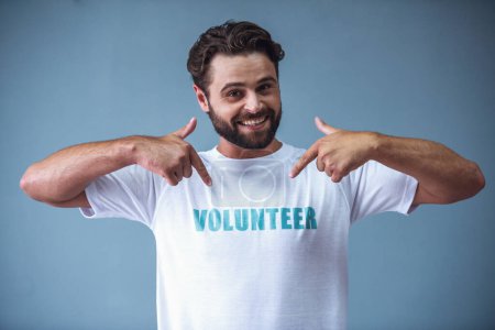 Photo for Handsome bearded man in volunteer t-shirt is pointing on inscription, looking at camera and smiling, on gray background - Royalty Free Image