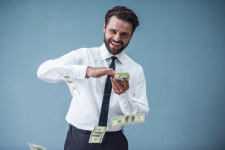 Foto de Hombre de negocios barbudo guapo en traje formal está lanzando billetes alrededor y sonriendo, sobre fondo gris - Imagen libre de derechos