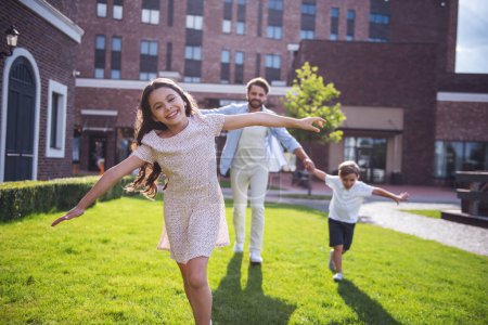 Téléchargez les photos : Des enfants joyeux et leur beau père barbu courent et sourient tout en jouant ensemble à l'extérieur - en image libre de droit