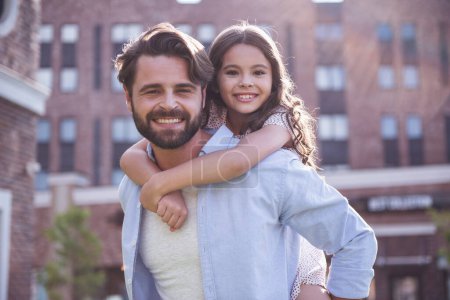 Photo for Handsome bearded dad and his cute little daughter are smiling while playing together outside, girl is sitting piggyback - Royalty Free Image