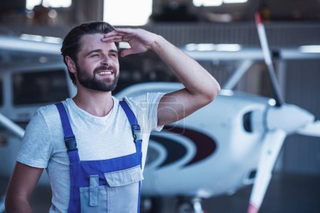 Foto de Guapo barbudo mecánico en uniforme está mirando hacia otro lado y sonriendo mientras está de pie cerca de un avión en el hangar - Imagen libre de derechos
