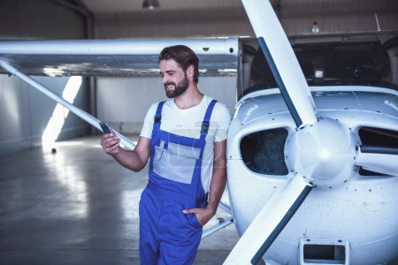 Photo for Handsome bearded mechanic in uniform is using a smart phone and smiling while leaning on the aircraft in hangar - Royalty Free Image