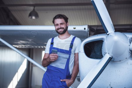 Foto de Guapo barbudo mecánico en uniforme está utilizando un teléfono inteligente y sonriendo mientras se apoya en el avión en el hangar - Imagen libre de derechos