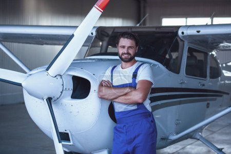 Foto de Guapo barbudo mecánico en uniforme está mirando a la cámara y sonriendo mientras se apoya en el avión en el hangar - Imagen libre de derechos