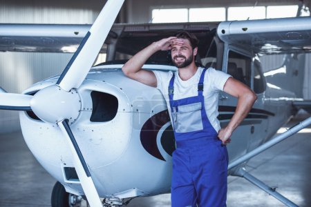 Foto de Guapo barbudo mecánico en uniforme está mirando hacia otro lado y sonriendo mientras se apoya en el avión en el hangar - Imagen libre de derechos