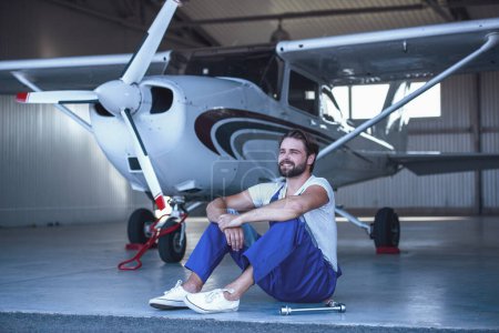 Foto de Mecánico barbudo guapo en uniforme mirando hacia otro lado y sentado en el piso del hangar cerca del avión - Imagen libre de derechos