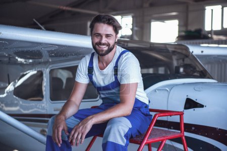 Foto de Guapo barbudo mecánico en uniforme está mirando a la cámara y sonriendo mientras está sentado cerca de la aeronave en el hangar - Imagen libre de derechos