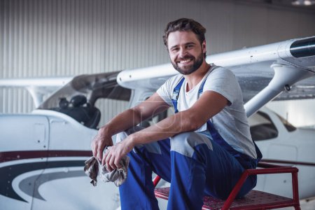 Foto de Guapo barbudo mecánico en uniforme está mirando a la cámara y sonriendo mientras está sentado cerca de la aeronave en el hangar - Imagen libre de derechos