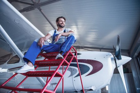 Foto de Vista de ángulo bajo del guapo mecánico barbudo en uniforme mirando hacia otro lado y sonriendo mientras está sentado cerca del avión en el hangar - Imagen libre de derechos