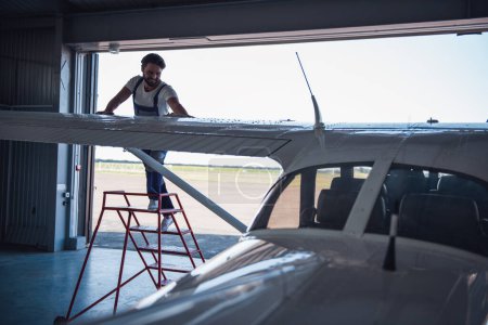 Foto de Guapo barbudo mecánico en uniforme sonríe mientras limpia el avión en el hangar - Imagen libre de derechos