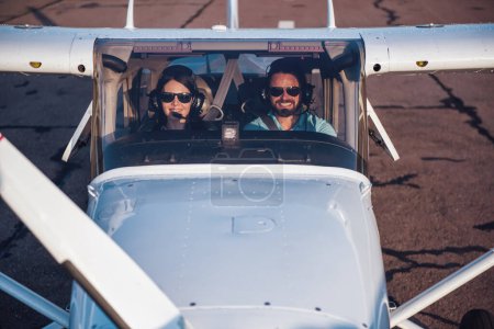 Photo for Beautiful couple in aviation headsets is looking at camera and smiling while sitting in aircraft ready to fly - Royalty Free Image