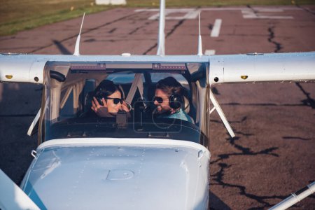 Foto de Hermosa pareja en auriculares de aviación está sonriendo mientras está sentado en los aviones, el hombre está ajustando los auriculares de la mujer - Imagen libre de derechos