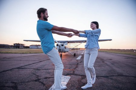 Photo for Beautiful romantic couple is holding hands, turning around and smiling on take-off ground near the aircraft - Royalty Free Image
