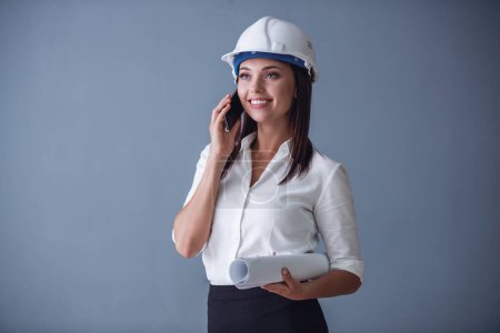 Photo for Beautiful young business woman in suit and protective helmet is talking on the mobile phone, holding rolled paper and smiling, on gray background - Royalty Free Image