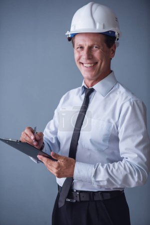 Photo for Handsome mature businessman in suit and protective helmet is making notes, looking at camera and smiling, on gray background - Royalty Free Image