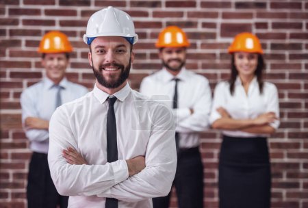 Photo for Handsome young bearded businessman in suit and protective helmet is looking at camera and smiling, his colleagues in the background - Royalty Free Image