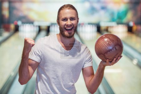 Téléchargez les photos : Beau jeune homme tient une balle de bowling, regardant la caméra et souriant prêt à jouer au bowling - en image libre de droit