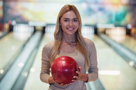 Téléchargez les photos : Jolie fille blonde tient une boule de bowling, regardant la caméra et souriant prêt à jouer au bowling - en image libre de droit