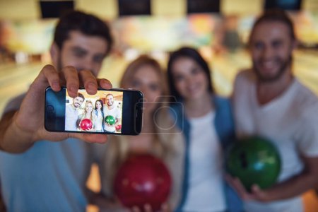 Foto de Felices jóvenes amigos están sosteniendo bolas, haciendo selfie y sonriendo mientras juegan bolos juntos, teléfono en foco - Imagen libre de derechos