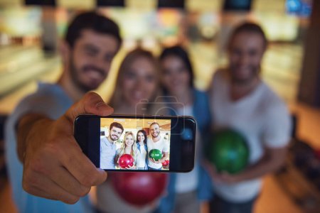 Foto de Felices jóvenes amigos están sosteniendo bolas, haciendo selfie y sonriendo mientras juegan bolos juntos, teléfono en foco - Imagen libre de derechos