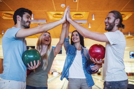 Téléchargez les photos : Heureux jeunes amis tiennent des boules, donnant cinq et souriant tout en jouant au bowling ensemble - en image libre de droit
