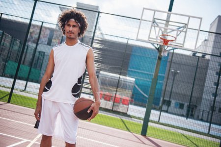 Photo for Attractive basketball player is holding a ball, looking away and smiling while standing on basketball court outdoors - Royalty Free Image