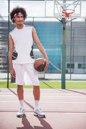 Téléchargez les photos : Pleine longueur de joueur de basket attrayant tenant une balle, regardant loin et souriant tout en se tenant sur le terrain de basket à l'extérieur - en image libre de droit