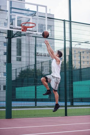 Foto de Guapo jugador de baloncesto está disparando una pelota a través de un aro mientras juega en la cancha de baloncesto al aire libre - Imagen libre de derechos
