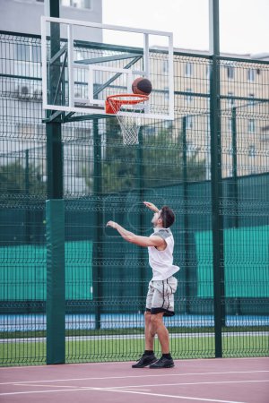 Téléchargez les photos : Beau joueur de basket-ball tire une balle à travers un cerceau tout en jouant sur le terrain de basket à l'extérieur - en image libre de droit