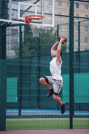 Téléchargez les photos : Beau joueur de basket-ball tire une balle à travers un cerceau tout en jouant sur le terrain de basket à l'extérieur - en image libre de droit