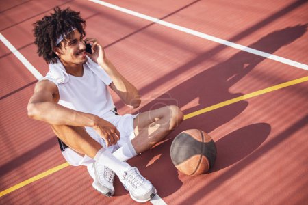 Téléchargez les photos : Joueur de basket attrayant parle sur le téléphone portable et souriant tout en étant assis sur le terrain de basket à l'extérieur - en image libre de droit