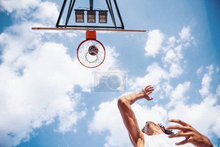 Téléchargez les photos : Vue du bas du joueur de basket tirant une balle à travers le cerceau tout en jouant sur le terrain de basket à l'extérieur - en image libre de droit