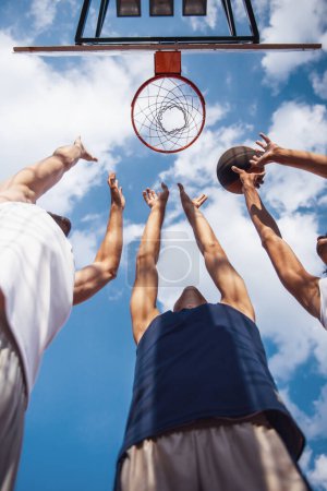 Foto de Vista inferior de jugadores de baloncesto guapos que disparan una pelota a través del aro mientras juegan en la cancha de baloncesto al aire libre - Imagen libre de derechos