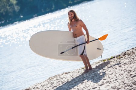 Téléchargez les photos : Beau jeune homme tient une planche et une pagaie et sourit tout en se tenant sur la plage - en image libre de droit