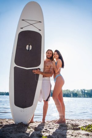 Téléchargez les photos : Beau jeune couple regarde la caméra et souriant tout en se tenant debout avec une planche à pagaie sur la plage - en image libre de droit