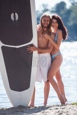 Téléchargez les photos : Beau jeune couple regarde la caméra, étreignant et souriant tout en se tenant debout avec une planche à pagaie sur la plage - en image libre de droit