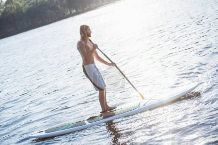 Téléchargez les photos : Beau jeune homme sourit tandis que SUP surf sur la rivière - en image libre de droit