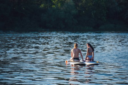 Photo for Beautiful young couple is talking and smiling while sitting on SUP boards on the river - Royalty Free Image