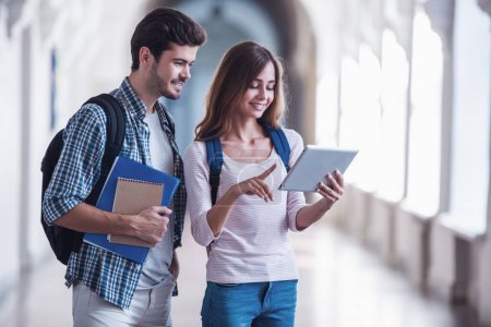 Téléchargez les photos : Jeunes étudiants attrayants utilisent une tablette numérique et souriant tout en se tenant dans la salle universitaire - en image libre de droit