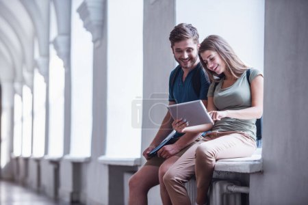 Foto de Atractivos estudiantes jóvenes están utilizando una tableta digital y sonriendo mientras están sentados en el alféizar de la ventana en el salón universitario - Imagen libre de derechos