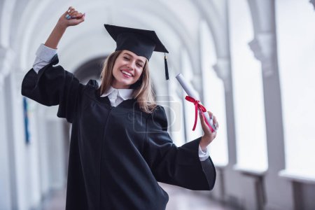 Foto de Atractiva joven graduada en vestido académico es la celebración de un diploma, mirando a la cámara y sonriendo mientras está de pie en la sala de la universidad - Imagen libre de derechos