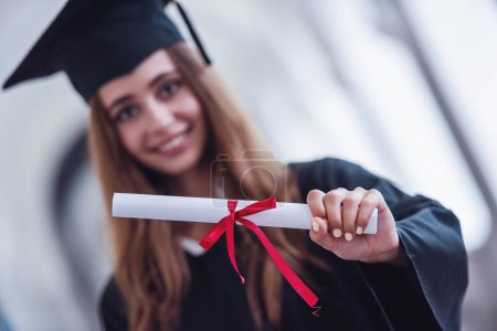 Photo for Attractive young female graduate in academic dress is holding a diploma, looking at camera and smiling while standing in university hall - Royalty Free Image