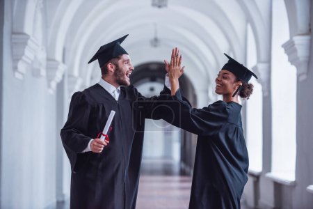 Photo for Happy young graduates in academic dresses are holding diplomas,  giving high five and smiling while standing in university hall - Royalty Free Image