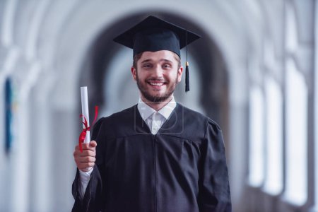 Téléchargez les photos : Beau jeune diplômé en tenue académique est titulaire d'un diplôme, regardant la caméra et souriant tout en se tenant dans la salle universitaire - en image libre de droit
