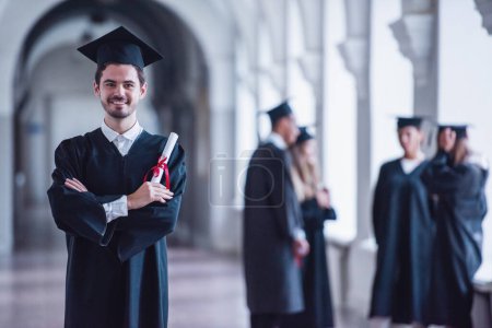Foto de Los graduados exitosos en vestidos académicos están hablando en el salón de la universidad, el tipo en primer plano está sosteniendo el diploma, mirando a la cámara y sonriendo - Imagen libre de derechos